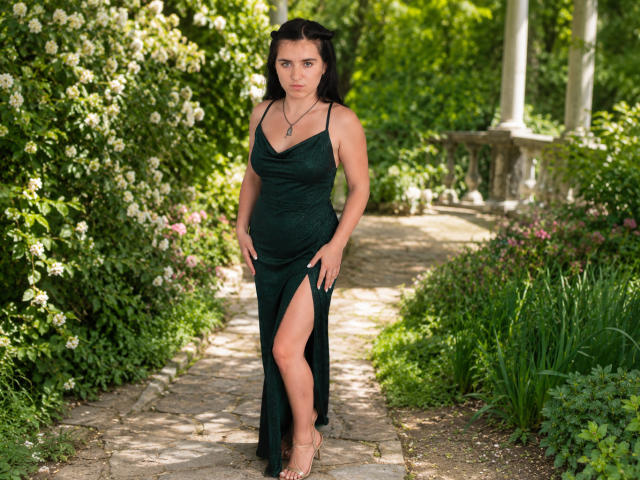 AnnaKimburli stands in a garden, wearing a dark green, slit-up dress, showing her tits and a leg, with a serious expression, black hair, green eyes, and a necklace.