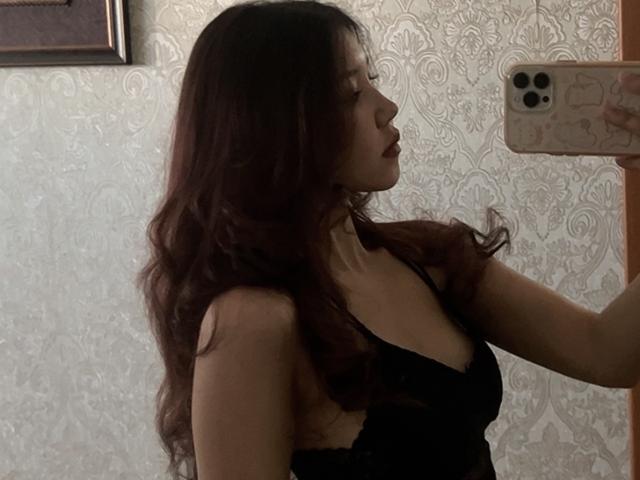 ElbaHuppenbauer, con i capelli castano ondulati, indossa biancheria intima di pizzo nero e sta scattando un selfie allo specchio, mostrando il seno nel riflesso, tenendo in mano un telefono rosa con tre fotocamere..