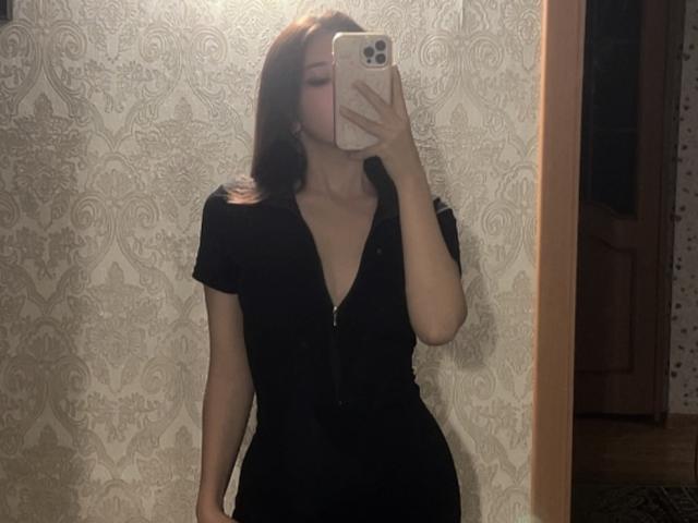 ElbaHuppenbauer trägt ein schwarzes Kleid mit tiefem V-Ausschnitt und kurzen Ärmeln und macht ein Selfie vor einem Spiegel mit ihrem Handy, wobei ihre Brüste zu sehen sind. Sie hat langes, braunes Haar und befindet sich in einem schlecht beleuchteten Raum..