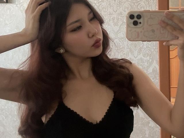 ElbaHuppenbauer mit dunklen, welligen Haaren, voller Lippen und einem schwarzen Spitzenoberteil, hält ein Telefon, macht ein Selfie im Spiegel und zeigt kleine Brüste und ein süßes Gesicht. Ein iPhone mit rosa Hülle in der Hand..