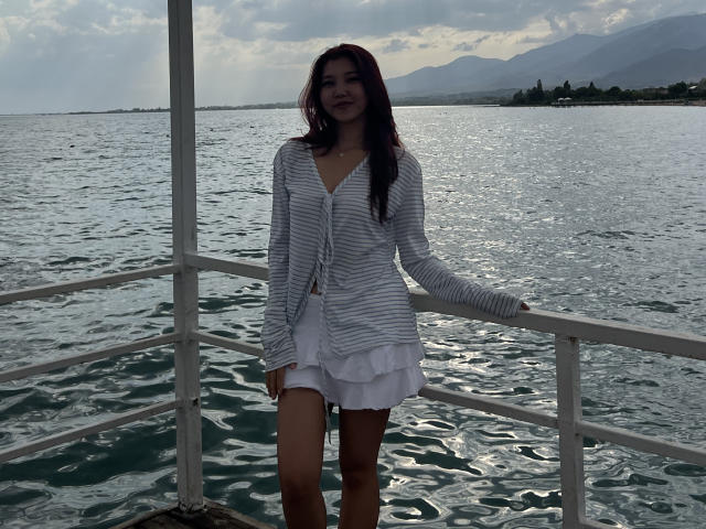 Elba Huppenbauer in piedi su una barca, indossa un cardigan bianco a righe e una gonna bianca con volant, ha lunghi capelli scuri e un leggero sorriso, con uno sfondo di acqua nuvolosa..