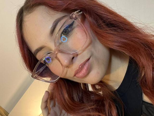 MiaaRouge, met rood haar, draagt een bril met heldere montuur en blauwe, ronde glazen, een zwarte top, heeft lichtroze lippen, een zachte glimlach en een lichte huid, en ze streelt haar haar..