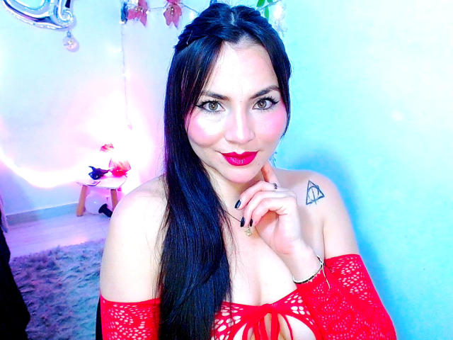 EmelySofia with long black hair, red lipstick, and off-shoulder red lace top, showing her tits, a triangle tattoo, and black nails, smiling in a blue-lit room.