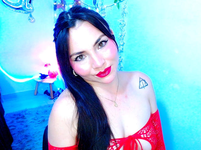 EmelySofia with long black hair, red off-shoulder dress, pink lipstick, tattoo on right shoulder, blue light, smiling, and blue background. Her tits are visible, she's hot.