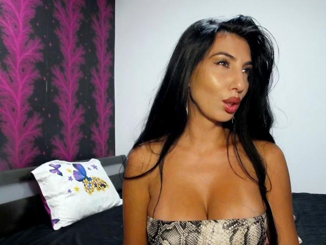 BrunetteBabe69, con lunghi capelli neri, labbra carnose e pelle abbronzata, indossa una maglietta con un motivo a serpente che mette in mostra il suo seno prosperoso. Sta guardando di lato con la bocca leggermente aperta..