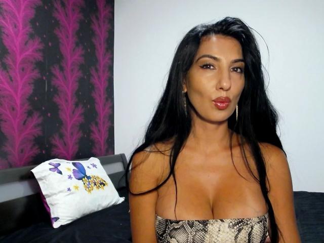 BrunetteBabe69 con lunghi capelli neri, pelle abbronzata e rossetto rosso, indossa un top senza spalline con motivo a serpente, mostrando il seno. Ha un'espressione leggermente accigliata, guardando la fotocamera..