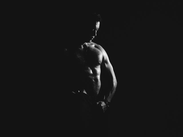 Il corpo muscoloso e senza camicia di AronWolff, con addominali e pettorali definiti, ombre scure che mettono in risalto la sua figura forte e scolpita, e una leggera vista di profilo, in una foto in bianco e nero ad alto contrasto..