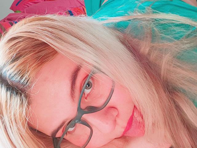 Akkanee has blonde hair, green glasses, and a pink nose, lying down with a green shirt, slightly smiling, looking at the camera, showing a cute face.