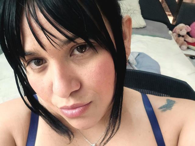 VivianaThompson has black hair, brown eyes, pink lips, light skin, small tattoo on right shoulder, wearing blue top, sitting in chair, looking at camera with slight smile.