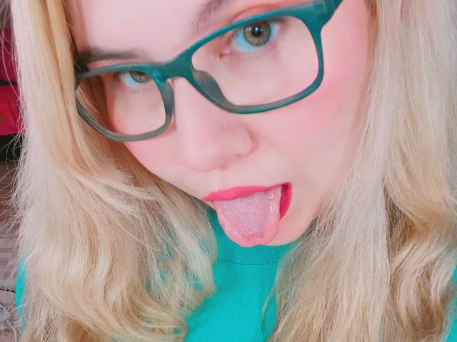 Akkanee with blonde hair, green glasses, and pink lipstick, sticking out a tongue with a pink, sparkling tongue piercing. She's wearing a teal shirt.