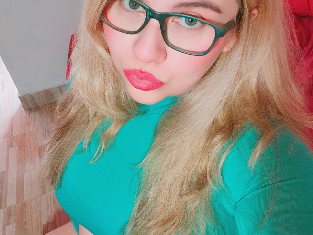 Akkanee with teal top, red lipstick, and green glasses, showing off her big tits, blonde hair, and pouty face, looking up at the camera.