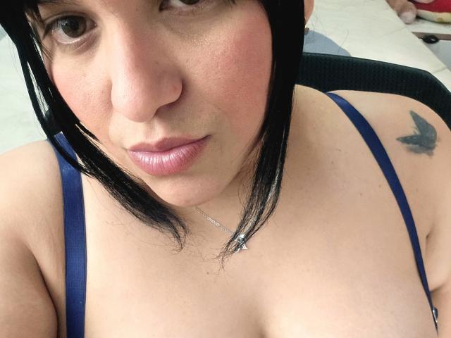 VivianaThompson has puffy pink lips, black hair, and a blue strap top showing her tits. She has a butterfly tattoo on her right shoulder and a silver necklace.