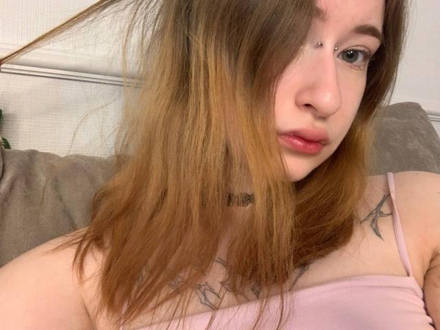 Marita Pavelko, mit blonden Haaren, Nasenpiercing und pinkem Lippenstift, trägt ein pinkes Oberteil, zeigt Tattoos auf ihrer Brust und eine Halskette, und sitzt auf einem grauen Sofa..