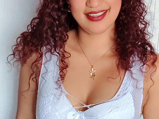 Sadieth with curly red hair, white lace top, and red lipstick, showing her tits and wearing a gold necklace with a flower pendant.