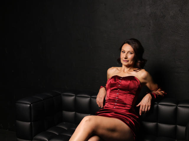 PinkSpark-hot with tits in a red satin dress, arms on black leather couch, legs crossed, dark brown hair, black wall, slight smile, sexy, slim, elegant, confident.