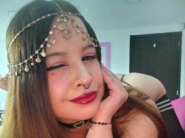 Melani Becker met rode lipstick, neusring en gouden hoofdketting, knipoogend, hand op haar wang, bruin haar, zwarte choker, roze oogschaduw, op een selfie, waarbij haar achterwerk op de achtergrond te zien is..
