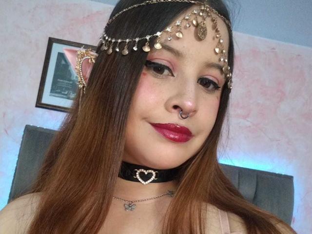 Melani Becker, met bruin haar, draagt een gouden hoofdtooi, een zwarte choker met een hartje, rode lippenstift en een neusring, en kijkt met een lichte glimlach in de camera. Haar borsten zijn gedeeltelijk zichtbaar..