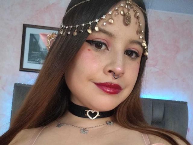 Melani Becker heeft bruin haar, rode lippenstift, een neusring, een zwarte choker met een hartje en een gouden hoofdband met hangende kralen. Ze kijkt naar de camera met een lichte glimlach..