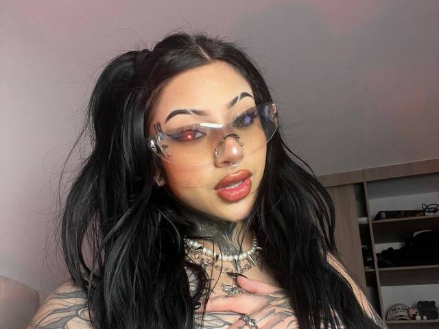 Milamonty with black pigtails, glasses, red and blue eyes, nose ring, and tattoos, wearing red lipstick, hand on chest, showing tits, in a selfie with pinkish light.