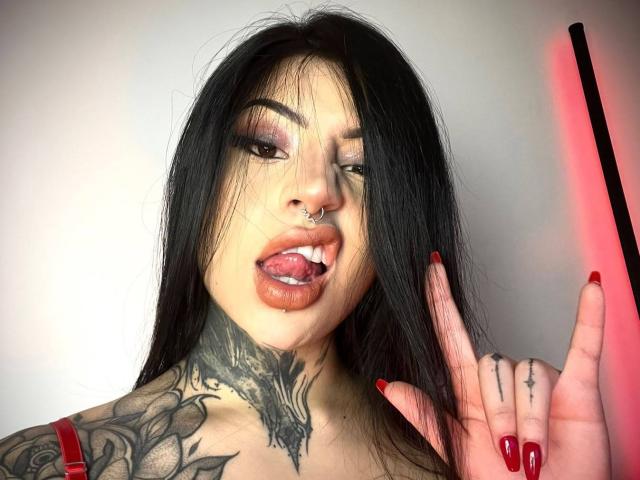 Milamonty with black hair, nose ring, and tattoos, sticking out tongue with red lipstick and nails, holding a red light. Visible tattoo on neck and hand tattoos.