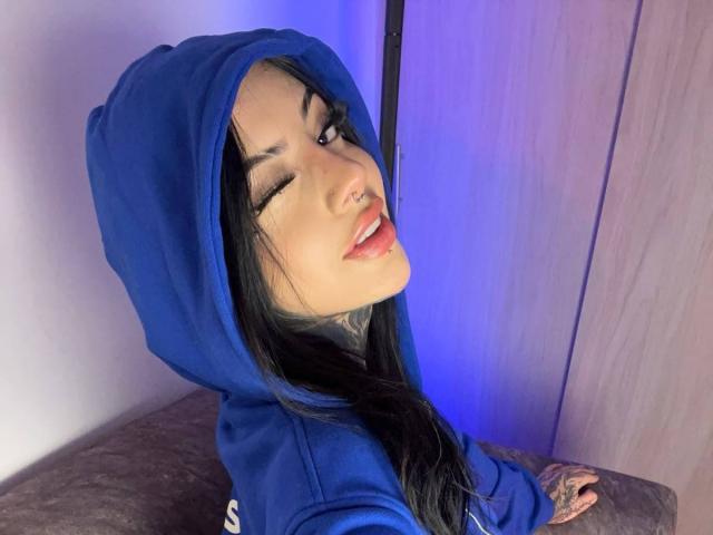 Milamonty with black hair, winking, wearing a blue hoodie, has tattoos on her hand, and a nose ring, sitting with her ass towards the camera.