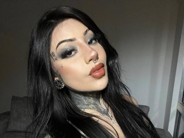 Milamonty has long black hair, dark makeup, tattoos, a nose ring, and a lip ring. She's looking at the camera with a pouty expression. Her tits are partially visible.