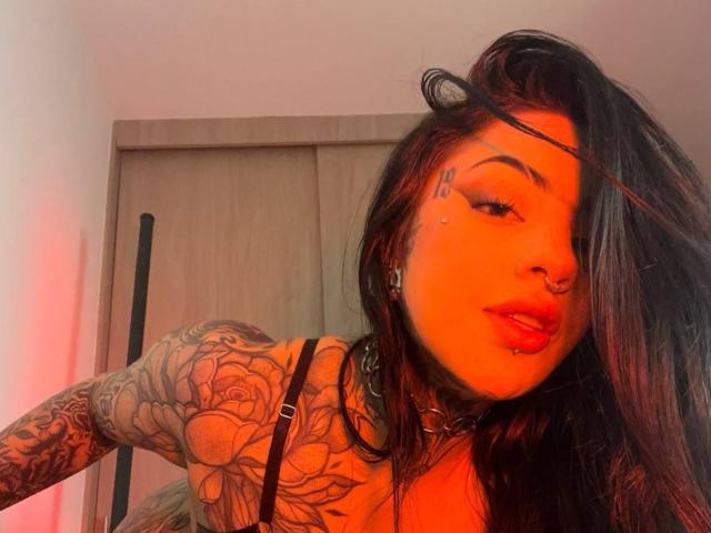 Milamonty with orange-tinted skin, black hair, tattoos on arm, nose ring, and lip piercing, wearing a black top, looking at camera with red lipstick.