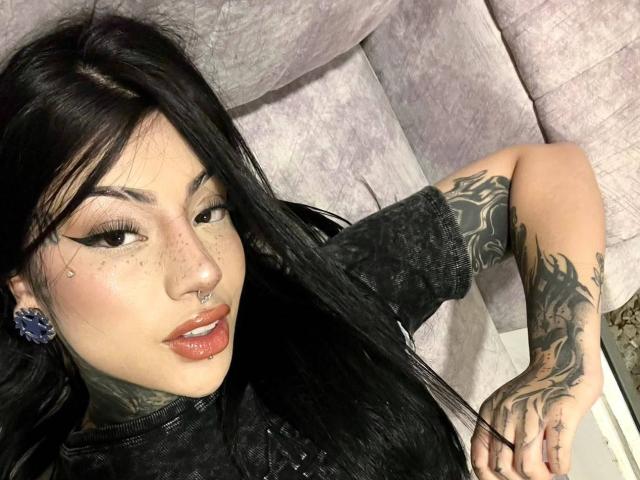 Milamonty with long black hair, red lips, tattoos, nose ring, and blue ear gauges, lying on a grey couch, showing off tattooed arm and black shirt.