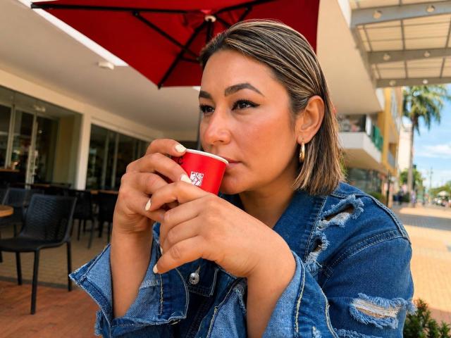 EmaFoxy, con piel morena, ojos marrones y cabello rubio hasta los hombros, bebe de una taza roja, vistiendo una chaqueta vaquera azul desgastada y aretes dorados, bajo un paraguas rojo..