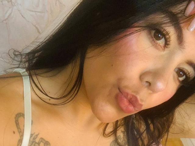 CatalinaReiyna with pouty lips, brown eyes, and black hair, wearing a white strap, has a tattoo on her left shoulder, and is looking at the camera.