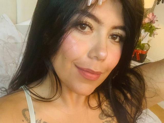 CatalinaReiyna has long black hair, brown eyes, and pink lips, with tattoos on her shoulder, wearing a white top, and a slight smile. Her hand is on her head.