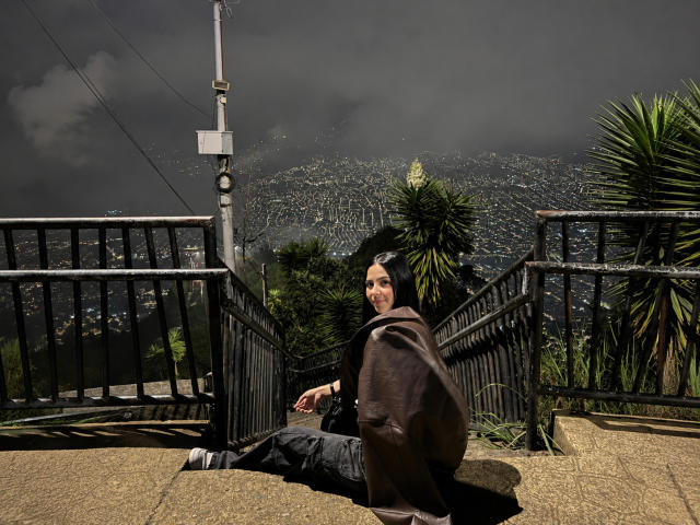 MiaAylen assise sur une bordure en béton, dos à la caméra, portant une veste marron, un pantalon noir et des chaussures grises, avec un palmier sur la droite, sous un ciel nuageux..