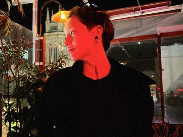 LissyLuxe with short red hair, black top, ear gauges, and sunglasses on head, looking to the side in red-lit night scene. Potted plant and street lamp in background.