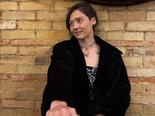 LissyLuxe has short brown hair, a black velvet coat, and a butterfly necklace, extending her hand forward with a slight smile. She has small tits and a cute face.