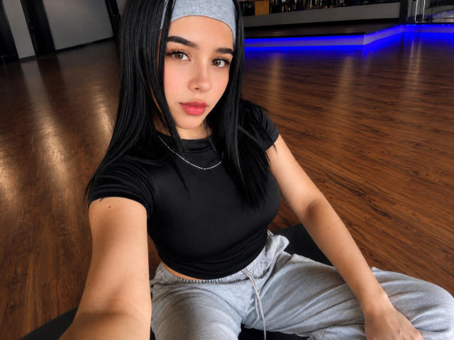 MiaAylen, avec de longs cheveux noirs, un bandeau gris, un haut noir, montrant de petites seins, un pantalon gris, assise sur un sol en bois, regardant la caméra avec une expression sérieuse, fond avec un éclairage néon bleu..