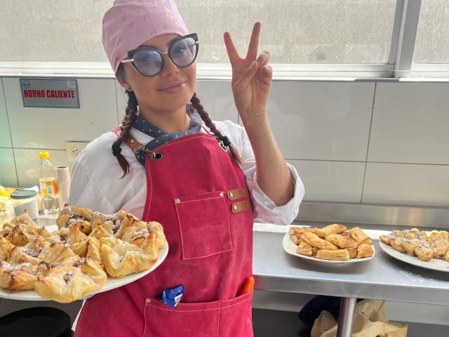 GalaMalkova con le trecce, che indossa un cappello rosa, un grembiule rosso, una camicia bianca e una sciarpa blu, tiene in mano un piatto di dolci sfogliati, fa un gesto di pace e indossa degli occhiali da sole alla moda..