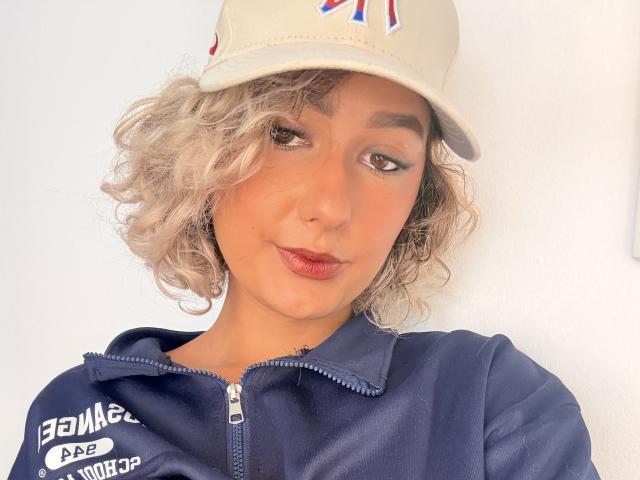 ElenaSaenz with curly blonde hair, brown eyes, and dark lipstick, wearing a beige cap and navy zip-up jacket, smiling softly. Her tits are small, and her ass is unseen.