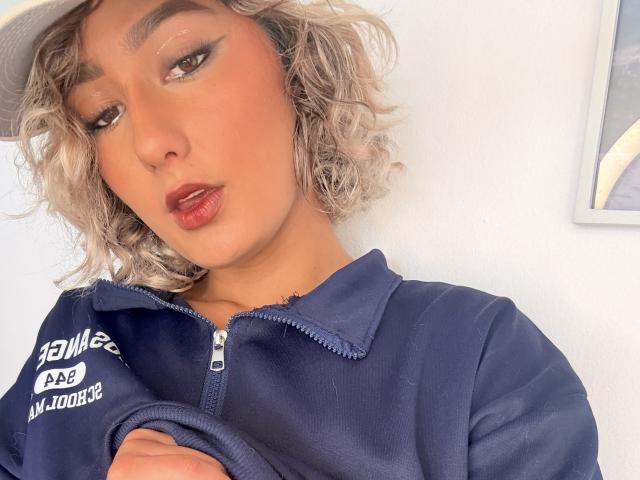ElenaSaenz with curly blonde hair, wearing a navy zip-up, has full red lips and subtle winged eyeliner, looking slightly to the side, wearing a white cap.