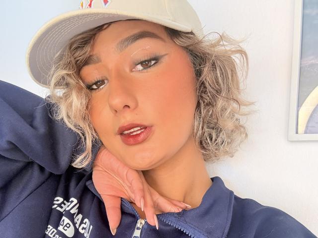 ElenaSaenz has curly blonde hair, a white cap, dark eyeliner, and is wearing a navy hoodie with her hand on her neck, showing off her glossy red lips.