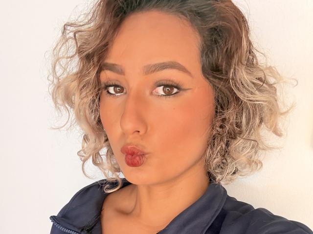 ElenaSaenz pouts her red lips, with curly blonde and brown hair, wearing a dark blue jacket, brown eyes looking slightly to the right.