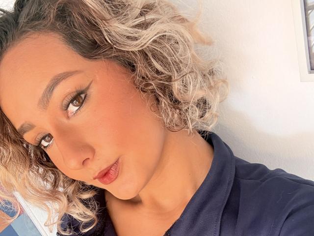 ElenaSaenz has curly blonde hair, brown eyes, and red lips, wearing a dark blue hoodie, with subtle winged eyeliner and a slight pout, in a bright, well-lit selfie.