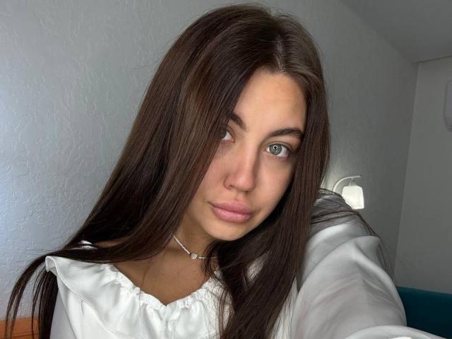 InesGueningsman with long brown hair, blue eyes, and pink lips, wearing a white shirt, has a slight smile, and is looking to the side in a selfie.