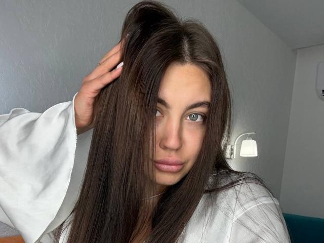 InesGueningsman with long brown hair, blue eyes, and pink lips, wearing a white robe, hand in hair, serious expression, soft light, white wall in background.