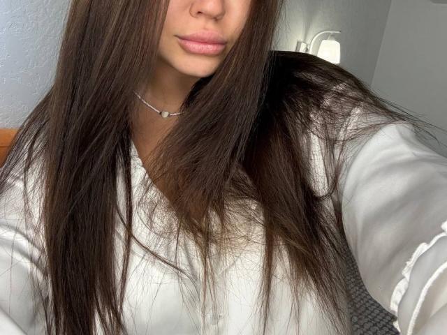 Ines Gueningsman, con lunghi capelli castani, una camicia bianca con bottoni, labbra rosate e una collana d'argento, sta scattando un selfie, mostrando leggermente il seno. I suoi capelli coprono la spalla destra..