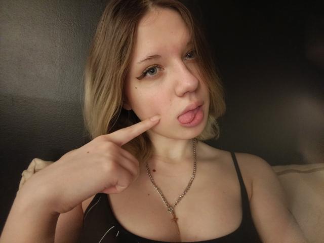 TameshaSchrimpf has blonde hair, pouty lips, and a small mole on her left cheek. She's sticking her tongue out and pointing at it with her finger. She's wearing a black top.