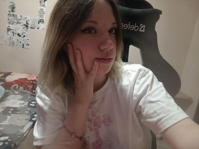 TameshaSchrimpf with pale skin, blue eyes, and blonde hair, wearing a white shirt, touching her cheek with her hand, sitting in a room with a black guitar.