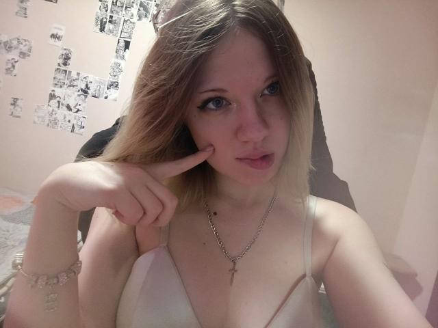 TameshaSchrimpf with blonde hair, blue eyes, and small tits, wearing a white bra, pointing at her slightly open mouth with her finger, necklace with cross, in a selfie.