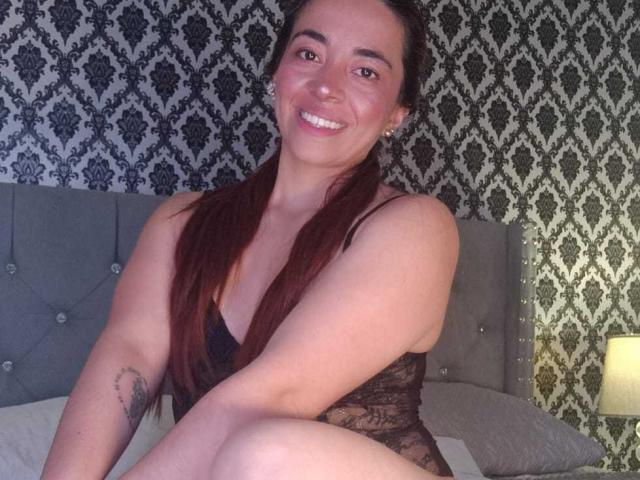 MadisonKoper, with long red hair, smiling, wearing a black lace bodysuit, showing a tattoo on her right arm, sitting on a bed with a grey headboard.