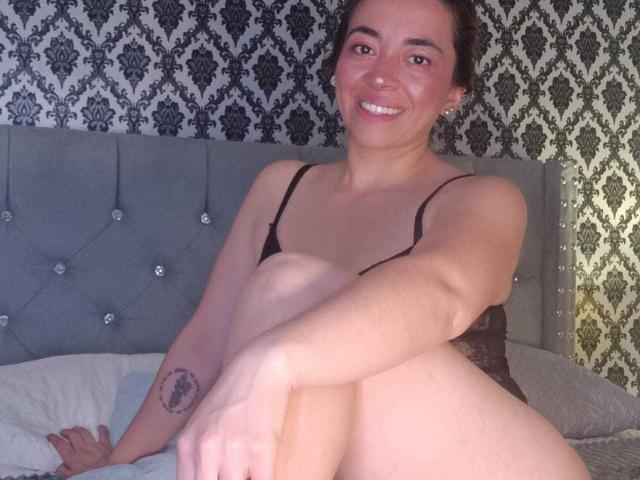 MadisonKoper sitting on a bed, smiling, wearing black lace lingerie, with her knees up, showing a tattoo on her right forearm, and her tits slightly visible.