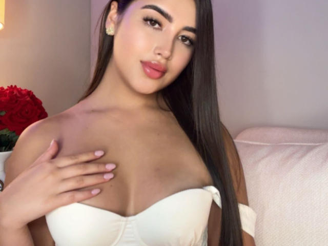AnnaPooky with long dark hair, wearing a white strapless top, touching her boobs with one hand, has pink lipstick, and is smiling slightly. Red roses in the background.